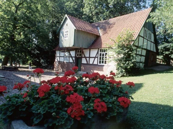 Fachwerkhaus mit roten Geranien im Vordergrund; umgeben von grünem Rasen und hohen Bäumen.