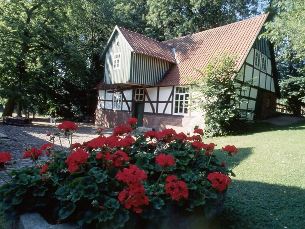Fachwerkhaus mit roten Geranien im Vordergrund; umgeben von grünem Rasen und hohen Bäumen.