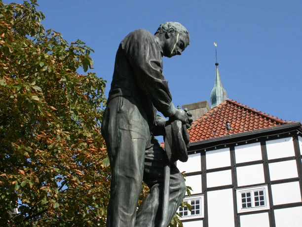 Bronzestatue eines Mannes, der eine Arbeitshaltung einnimmt, vor einem Fachwerkgebäude und blauem Himmel.