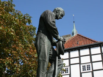 image Bronzestatue eines Mannes, der eine Arbeitshaltung einnimmt, vor einem Fachwerkgebäude und blauem Himmel.