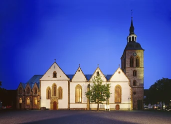 Gotische Kirche im Abendlicht, mit beleuchteten Fenstern und einem markanten Turm vor blauem Himmel.