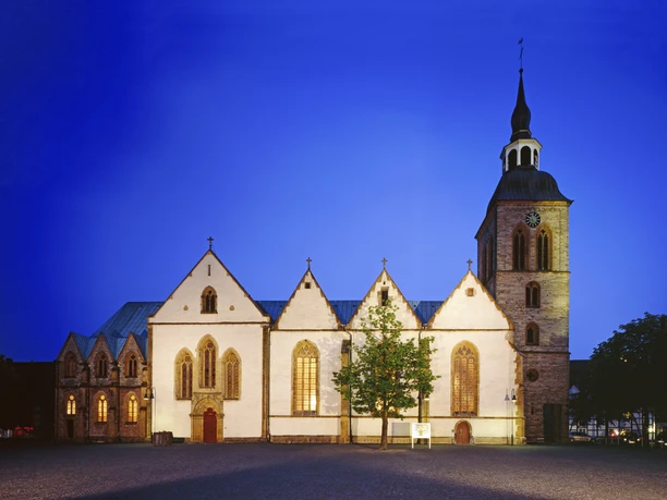 Gotische Kirche im Abendlicht, mit beleuchteten Fenstern und einem markanten Turm vor blauem Himmel.