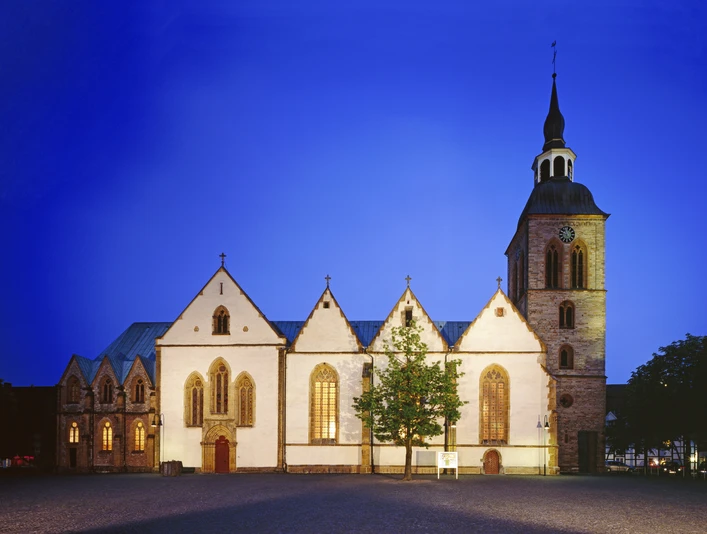 Gotische Kirche im Abendlicht, mit beleuchteten Fenstern und einem markanten Turm vor blauem Himmel.