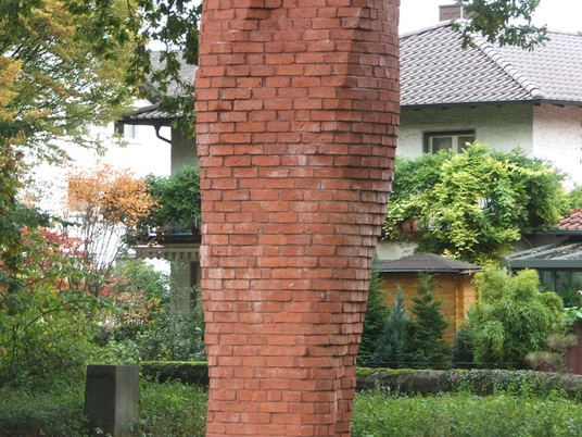 Backsteinstele im Park von Bäumen umgeben, symbolisiert Reflexion und Ruhe im öffentlichen Raum.
