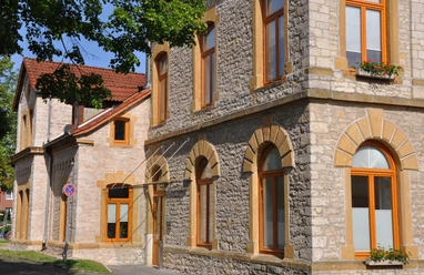 Steinernes Rathausgebäude mit großen Fenstern und rötlichem Rahmen, umgeben von Bäumen im Sonnenschein.