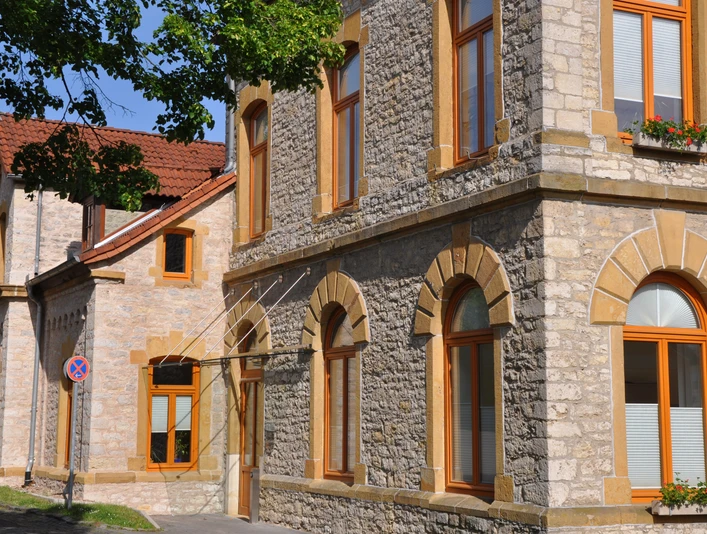 Steinernes Rathausgebäude mit großen Fenstern und rötlichem Rahmen, umgeben von Bäumen im Sonnenschein.