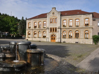 Historisches Gebäude aus Naturstein mit rotem Dach hinter einem modernen, plätschernden Brunnen.