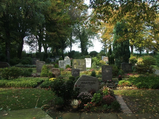 Friedhof Stapelage Eine ruhige Gedenkstätte mit gepflegten Gräbern, umgeben von hohen Bäumen und buntem Laub.