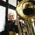 Musiker von nah und fern genießen die freundliche Atmosphäre im Jazzkotten Älterer Mann mit grauem Haar spielt eine glänzende Tuba und lächelt, traditionelle Fachwerkfassade im Hintergrund.