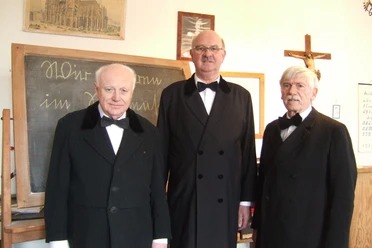 Drei ältere Herren in schwarzen Anzügen im Dorfschulmuseum vor einer Schultafel.