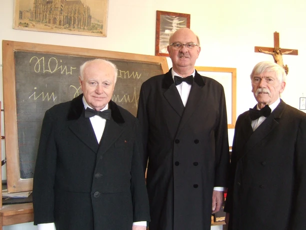Dorfschullehrer im Dorfschulmuseum Drei ältere Herren in schwarzen Anzügen im Dorfschulmuseum vor einer Schultafel.