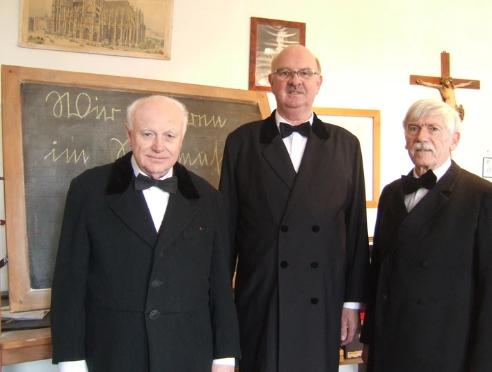 Dorfschullehrer im Dorfschulmuseum Drei ältere Herren in schwarzen Anzügen im Dorfschulmuseum vor einer Schultafel.