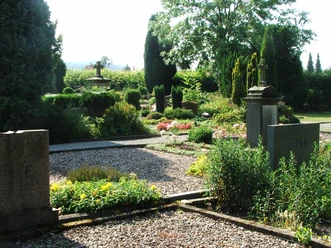 Grünbewachsener Friedhof mit gepflegten Grabsteinen und Wegen, umgeben von Bäumen und Gebüschen.