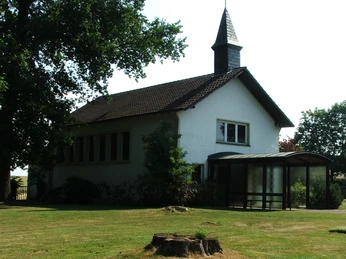 Friedhofskapelle Heiden mit dunklem Dach und kleinem Turm, umgeben von grünen Bäumen und Wiesen.