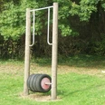 Spielstation Hiddentrup Ein Spielgerät mit Reifenrolle auf einem Rasenplatz, umgeben von dichter grüner Vegetation.