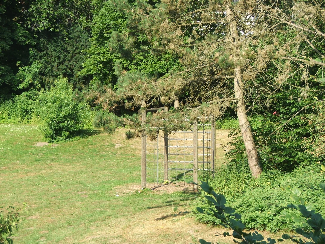 Spielplatz Haus des Gastes Klettergerüst aus Holz inmitten eines grünen Parks mit dichten Bäumen und Schattenbereichen.