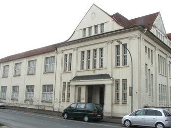 Historische, zweistöckige Fassade mit Giebel, großen Fenstern und geparkten Autos an der Straße.