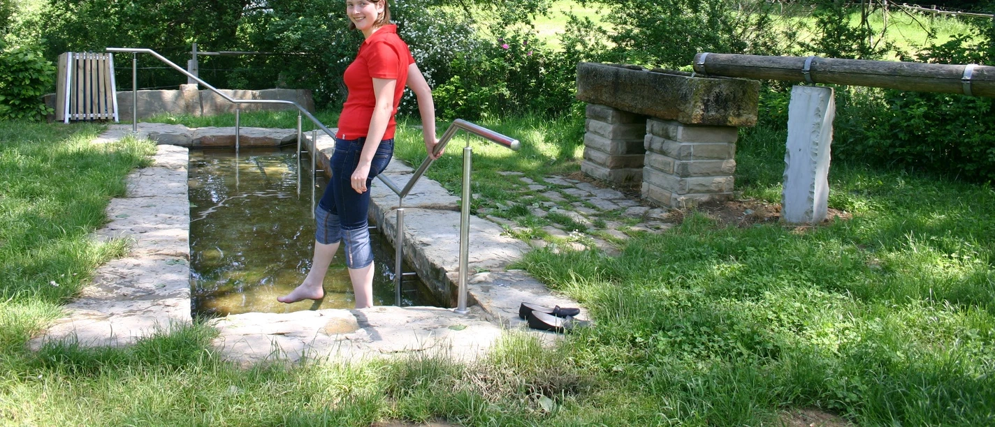 Eine Frau nutzt die Kneipp-Anlage im Golmeketal, umgeben von üppigem Grün und einem klaren Himmel.