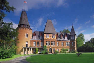 Schloss Hollwinkel mit markanten Türmen und Fachwerkgebäude umgeben von gepflegter Grünanlage.