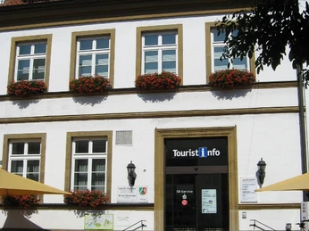 Touristikinformation Zweistöckiges Gebäude mit Tourist-Info, weißer Fassade, Blumen an Fenstern und gelben Sonnenschirmen.