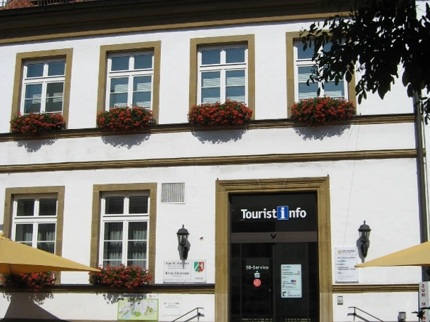 Touristikinformation Zweistöckiges Gebäude mit Tourist-Info, weißer Fassade, Blumen an Fenstern und gelben Sonnenschirmen.