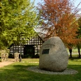 Großer Findling mit Gedenktafel, umgeben von Bäumen, befestigter Weg führt zu einem Fachwerkhaus.