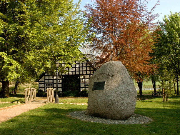Frontansicht vom Haus der Begegnung Großer Findling mit Gedenktafel, umgeben von Bäumen, befestigter Weg führt zu einem Fachwerkhaus.