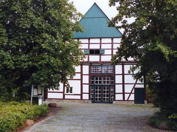 Domhof©Stadt Rheda-Wiedenbrück.jpeg Fachwerkhaus mit grünem Giebeldach, umgeben von Bäumen, steht auf gepflastertem Platz.