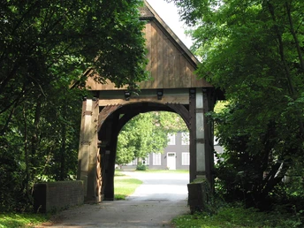 Gut Schledebrück Holztor mit traditionellem Design, eingerahmt von lebendigem, grünem Laub. Weg führt hindurch.