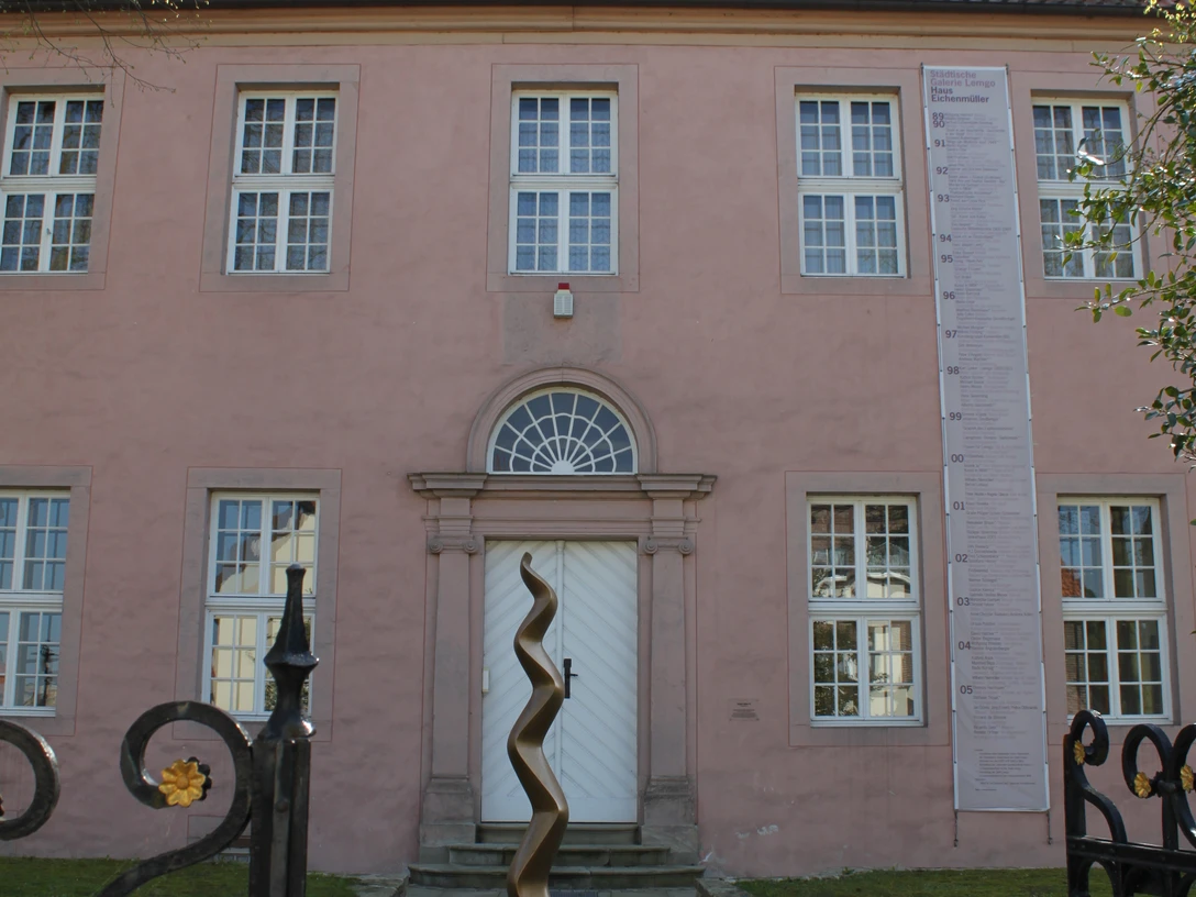 Städtische Galerie Eichenmüllerhaus Ehemaliges Wohnhaus mit rosa Fassade und modernen Kunstskulpturen vor dem Eingang, umgeben von Bäumen.