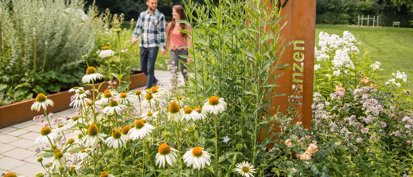 Zwei Personen spazieren durch eine gepflegte Gartenanlage mit vielfältigen Kräuter- und Blumenbeeten.