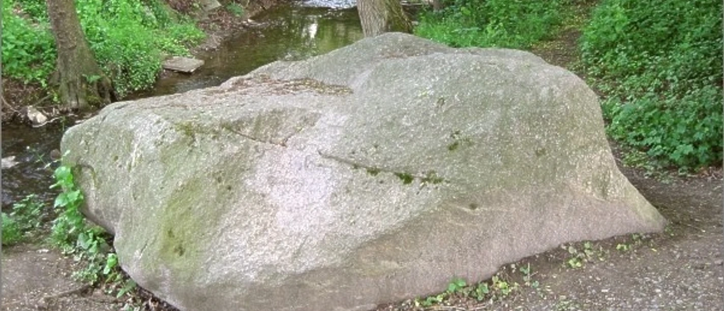 Eselstein Großer, flacher Findling an einem Bach in bewaldeter Umgebung, umgeben von Kies und Bäumen.