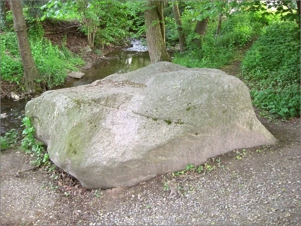 Eselstein Großer, flacher Findling an einem Bach in bewaldeter Umgebung, umgeben von Kies und Bäumen.