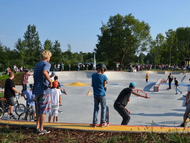 Skatepark "Altes Klärwerk" in Rheda-Wiedenbrück, Bereich Mittelhegge Jugendliche und Kinder nutzen einen belebten Skatepark bei sonnigem Wetter, umgeben von Bäumen.