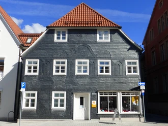 Traditionelles dreistöckiges Gebäude mit Schieferfassade und roten Ziegeldach, umgeben von modernen Bauten.
