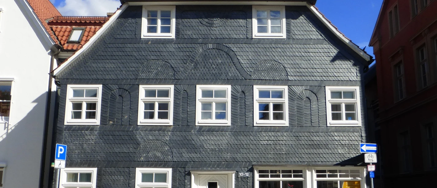 Traditionelles dreistöckiges Gebäude mit Schieferfassade und roten Ziegeldach, umgeben von modernen Bauten.