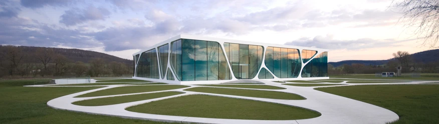Modernes Gebäude mit großen Glasflächen und geschwungener Architektur in einer grünen Landschaft.