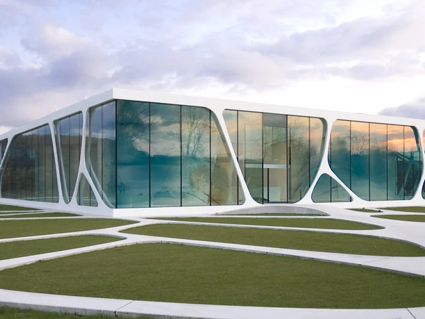 Modernes Gebäude mit großen Glasflächen und geschwungener Architektur in einer grünen Landschaft.