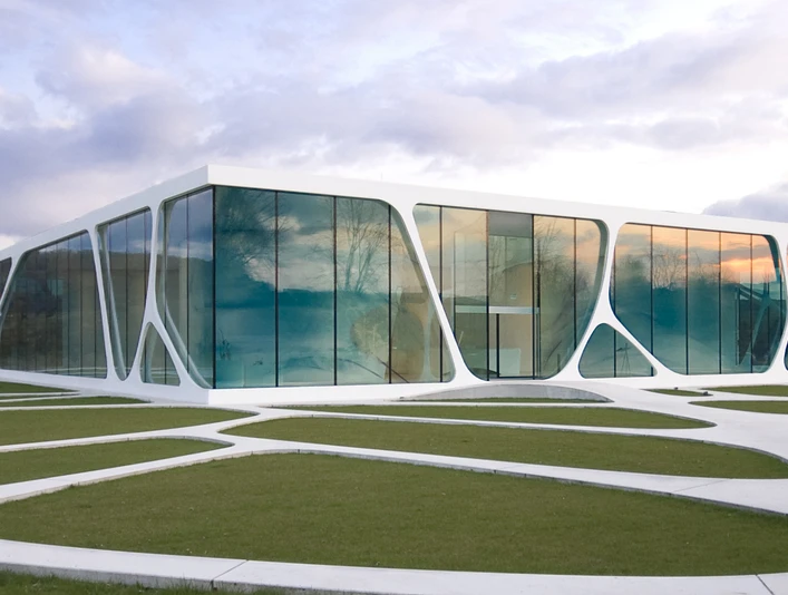 Modernes Gebäude mit großen Glasflächen und geschwungener Architektur in einer grünen Landschaft.