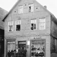 Historisches Fachwerkhaus mit Menschen an Fenstern und Eingang, Schaufenster mit Hüten und Waren.