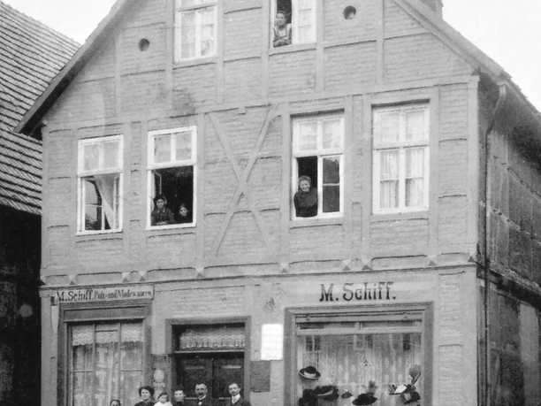 Historisches Fachwerkhaus mit Menschen an Fenstern und Eingang, Schaufenster mit Hüten und Waren.