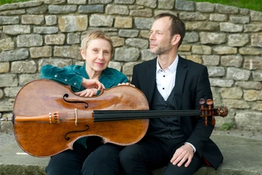 WEGE ERLEBEN Ein Mann und eine Frau sitzen vor einer Steinmauer, die Frau hält ein Cello in der Hand.