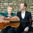 WEGE ERLEBEN Ein Mann und eine Frau sitzen vor einer Steinmauer, die Frau hält ein Cello in der Hand.