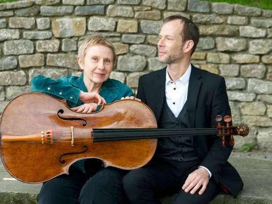 WEGE ERLEBEN Ein Mann und eine Frau sitzen vor einer Steinmauer, die Frau hält ein Cello in der Hand.