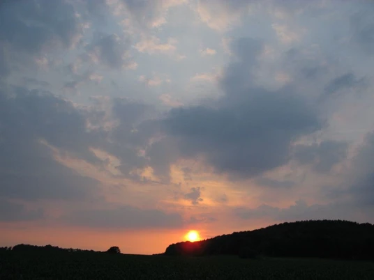 Sonnenuntergang Orange-roter Sonnenuntergang über bewaldetem Hügel, umgeben von dichten, grauen Wolkenformationen.