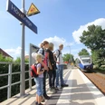 Eine Familie wartet am Bahnhof Hövelriege auf den einfahrenden Zug bei strahlendem Sonnenschein.