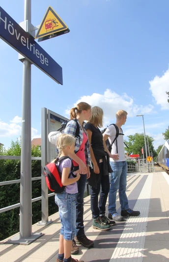 Eine Familie wartet am Bahnhof Hövelriege auf den einfahrenden Zug bei strahlendem Sonnenschein.