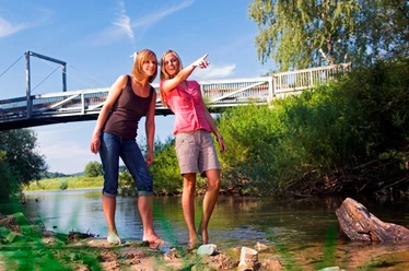 Erfrischung im Reich der Prachtlibellen Zwei Frauen barfuß in einem Fluss, eine zeigt lächelnd auf eine Brücke an einem sonnigen Tag.