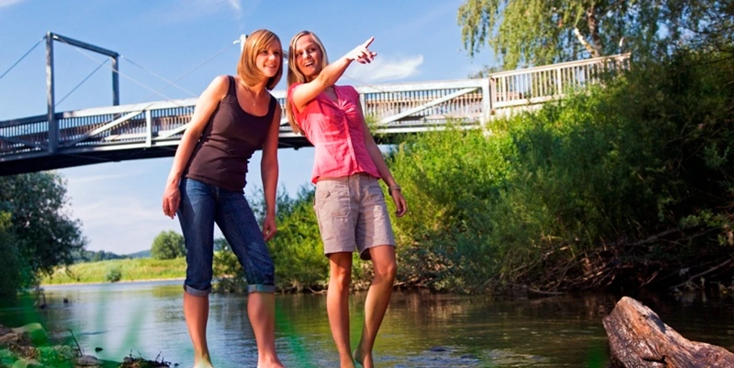 Erfrischung im Reich der Prachtlibellen Zwei Frauen barfuß in einem Fluss, eine zeigt lächelnd auf eine Brücke an einem sonnigen Tag.