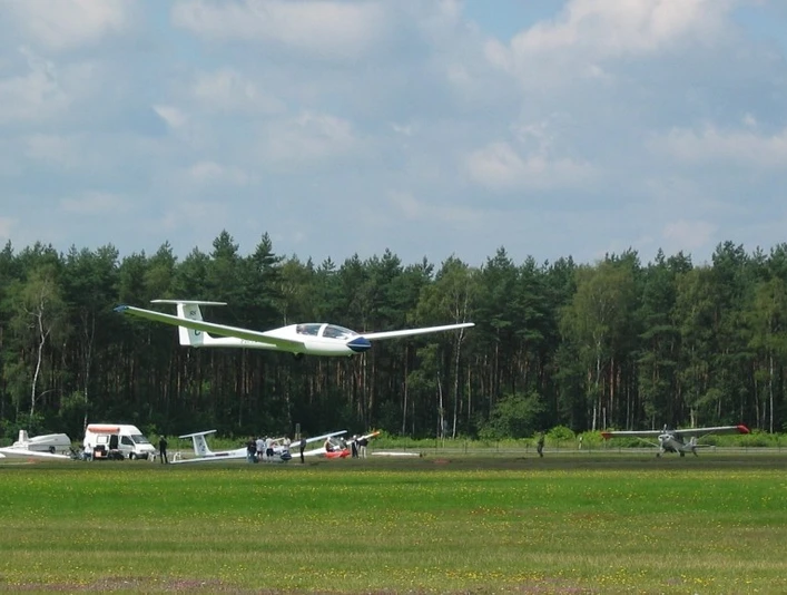 Segelflugzeug hebt vom grünen Flugplatz ab, im Hintergrund dichter Wald und ruhiger Himmel.